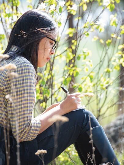 A person writes on a notebook while sitting amongst trees