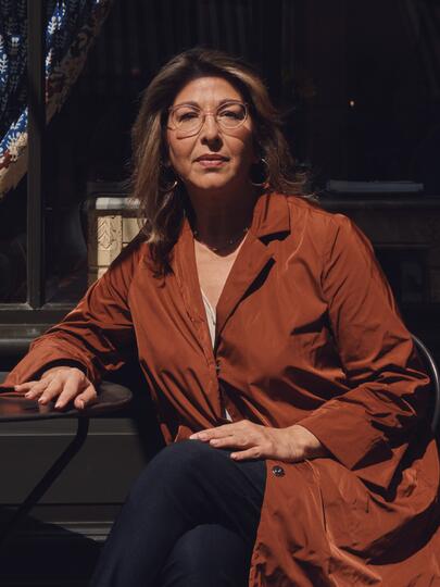 A photo of Naomi Klein seated at a metal table against a dark background, wearing glasses and a russet overcoat.