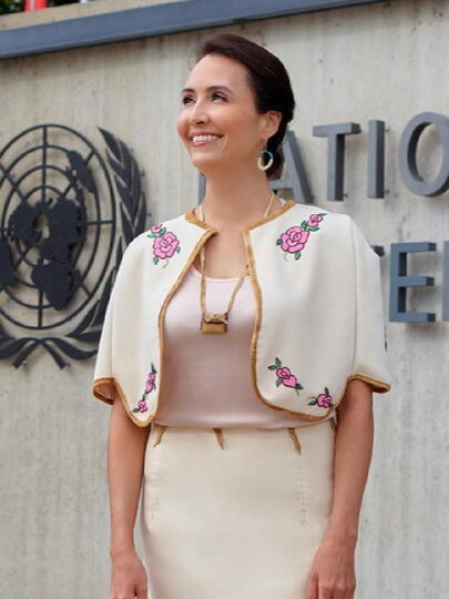 Standing in front of a United Nations sign, Jennifer Duncan wears a white blazer and skirt with rose designs and a gold trim.