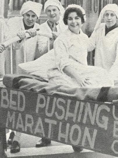  Four people standing around a person in a bed. Attached to the bed is a sign that reads: "UBC Bed Pushing Marathon"