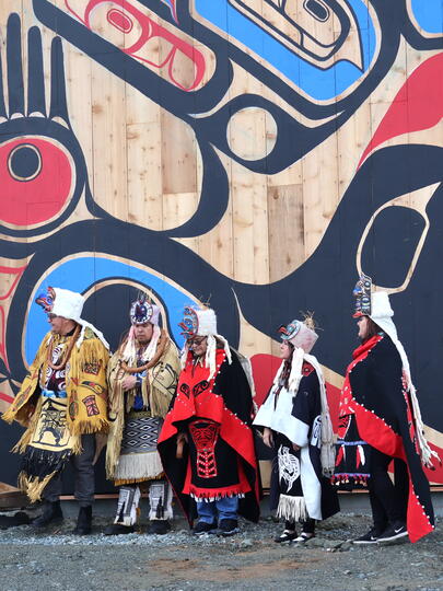 Five people in Indigenous outfits in front of wall with Indigenous artwork